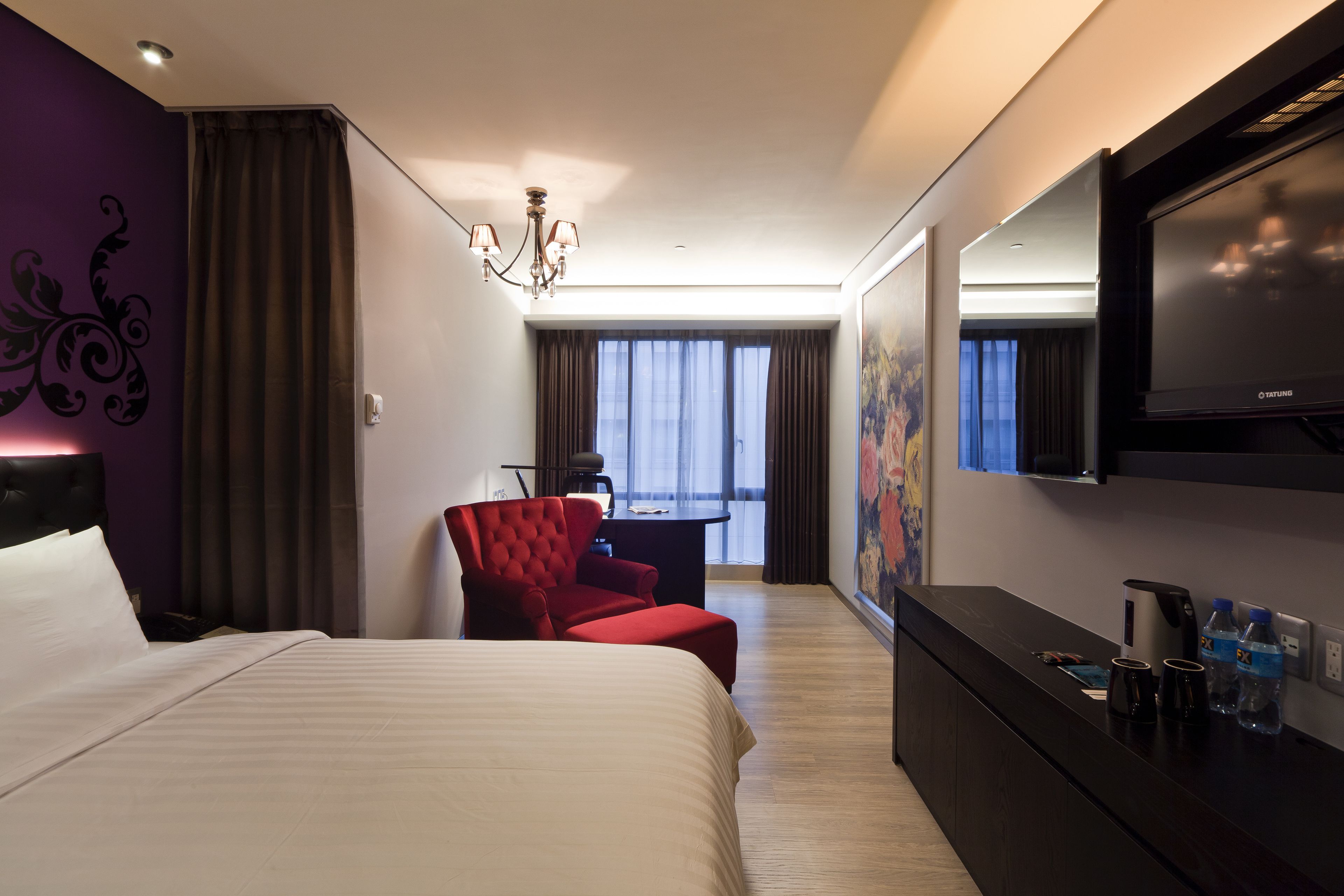 A hotel room with a large bed, a red armchair, a desk with a chair, a flat-screen TV, and a painting on the wall.
