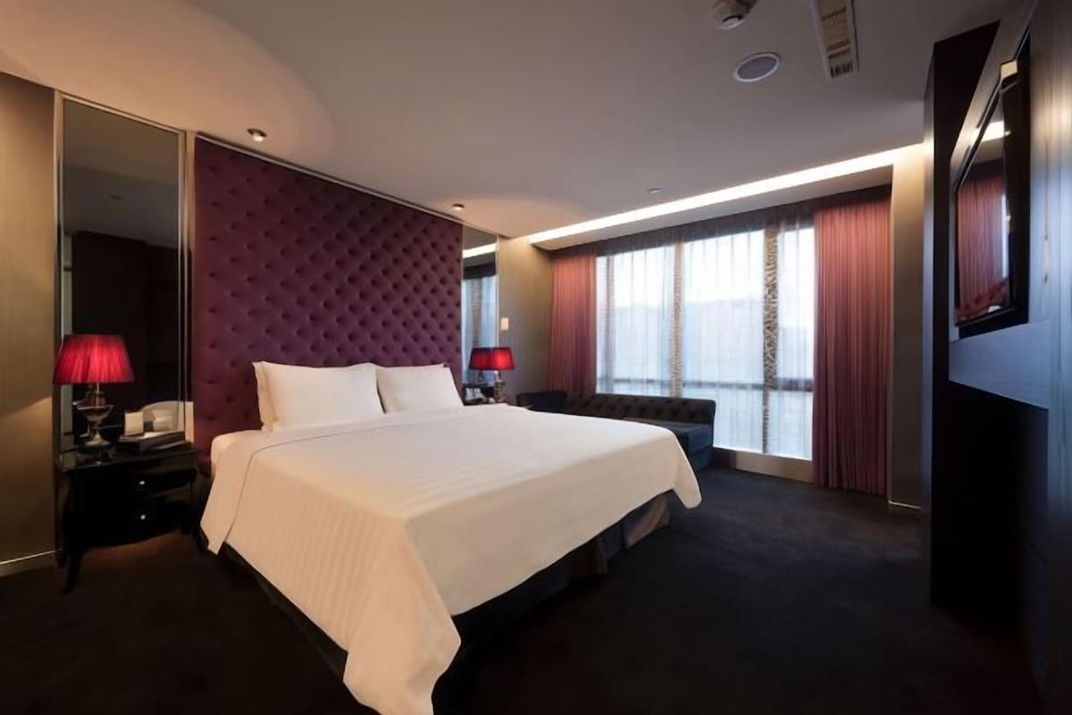 A hotel room with a large bed, a flat-screen TV, a desk with a chair, and a large window with curtains.