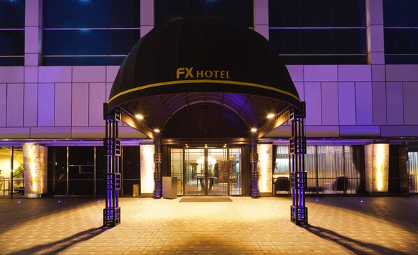 La entrada del Hotel FX, con un diseño moderno, que presenta una gran estructura en forma de cúpula y pilares iluminados.