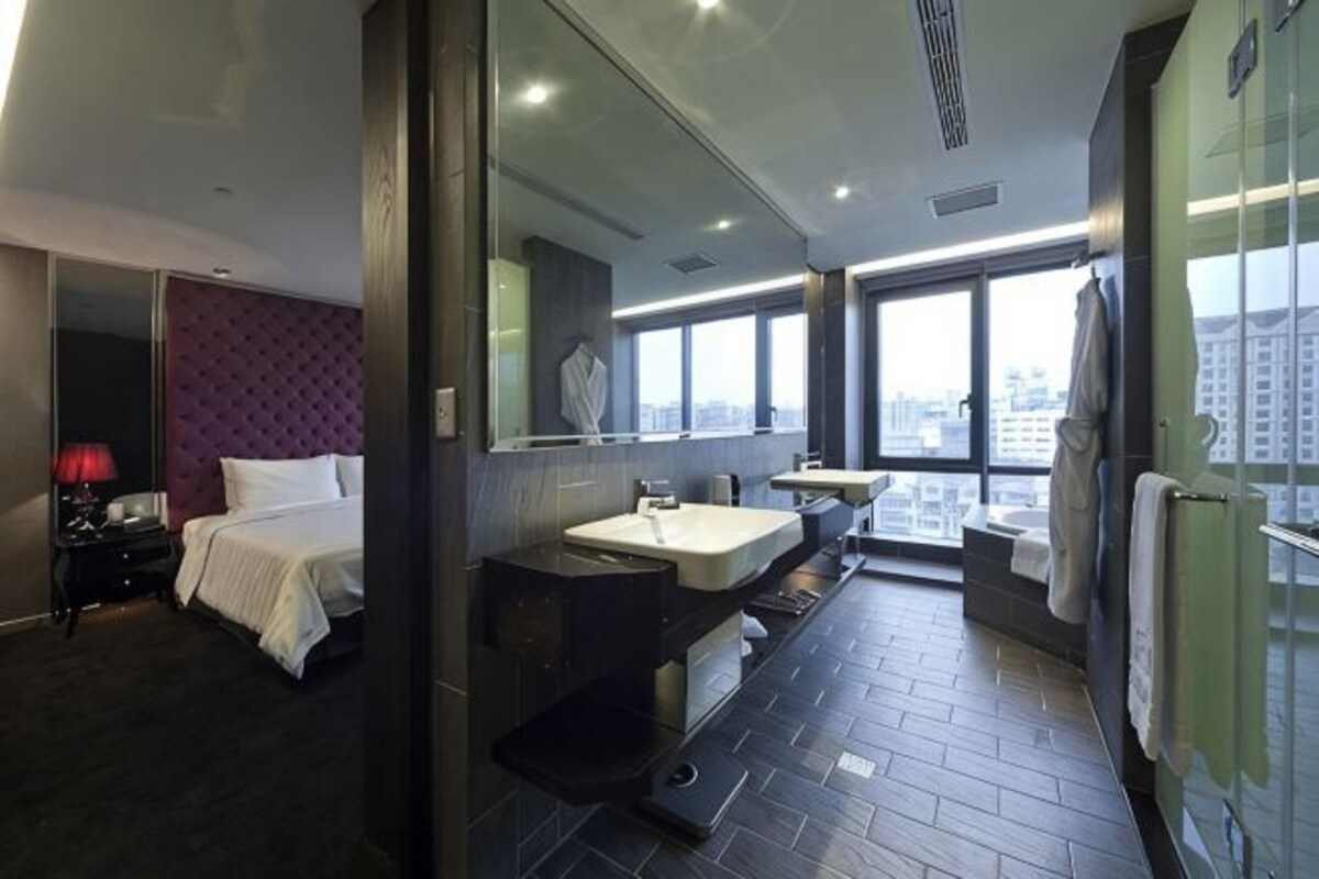 Una habitación de hotel moderna con una cama, un espejo grande, un baño con lavamanos e inodoro, y vistas de la ciudad a través de amplias ventanas.