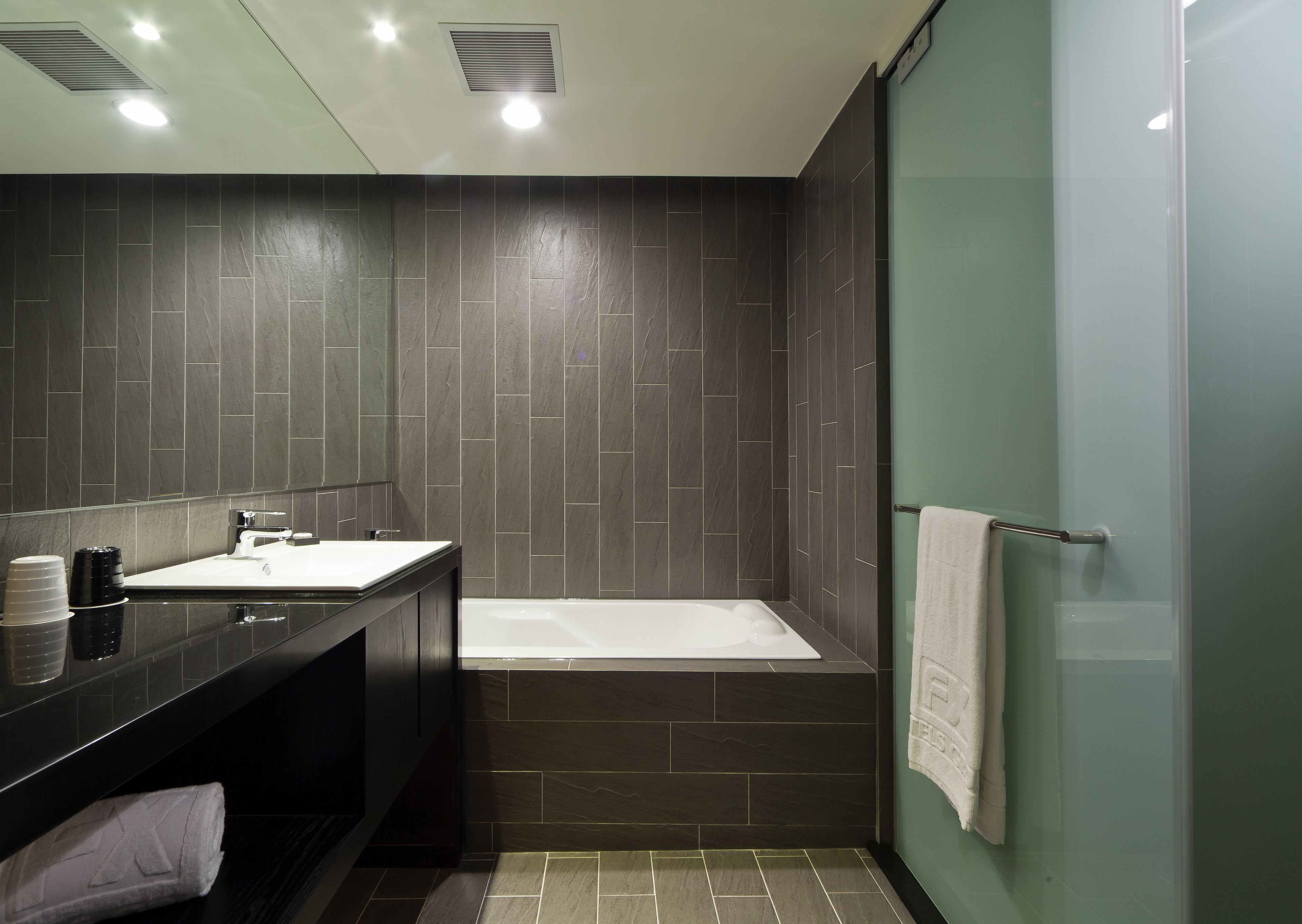 A modern bathroom with a dark tiled wall, a white sink, a bathtub, and a glass shower enclosure.