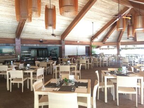 A spacious restaurant with high ceilings and wooden beams, featuring numerous tables and chairs, a kitchen area, and hanging pendant lights. - Bluewater Panglao Beach Resort (Panglao)