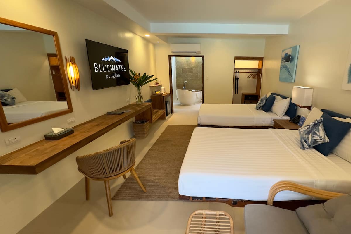 Habitación de hotel con dos camas, un escritorio, una silla, un televisor y un baño visible a través de una puerta abierta.