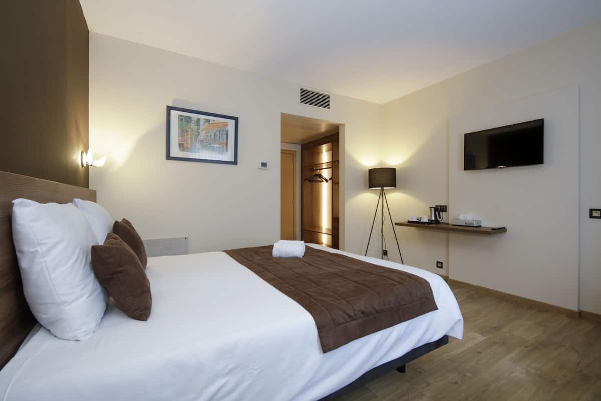 Une chambre d’hôtel avec un grand lit, un téléviseur à écran plat fixé au mur, une table de chevet avec une lampe et un tableau encadré au mur.