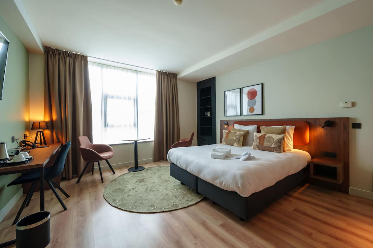 Une chambre d’hôtel moderne avec un grand lit, un bureau avec une lampe, une chaise et une vue sur la ville par la fenêtre.