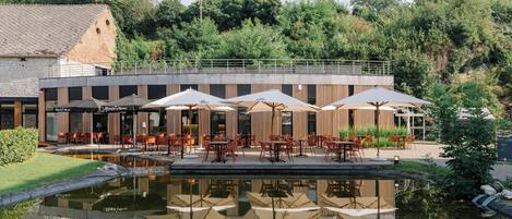 A restaurant with outdoor seating by a pond, featuring umbrellas and wooden tables.