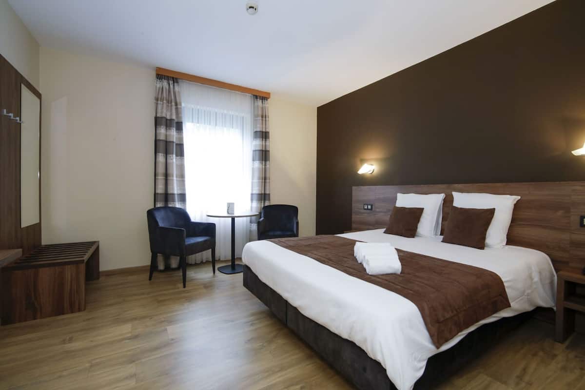 Une chambre d’hôtel avec un grand lit, deux chaises, une petite table et une fenêtre avec des rideaux.