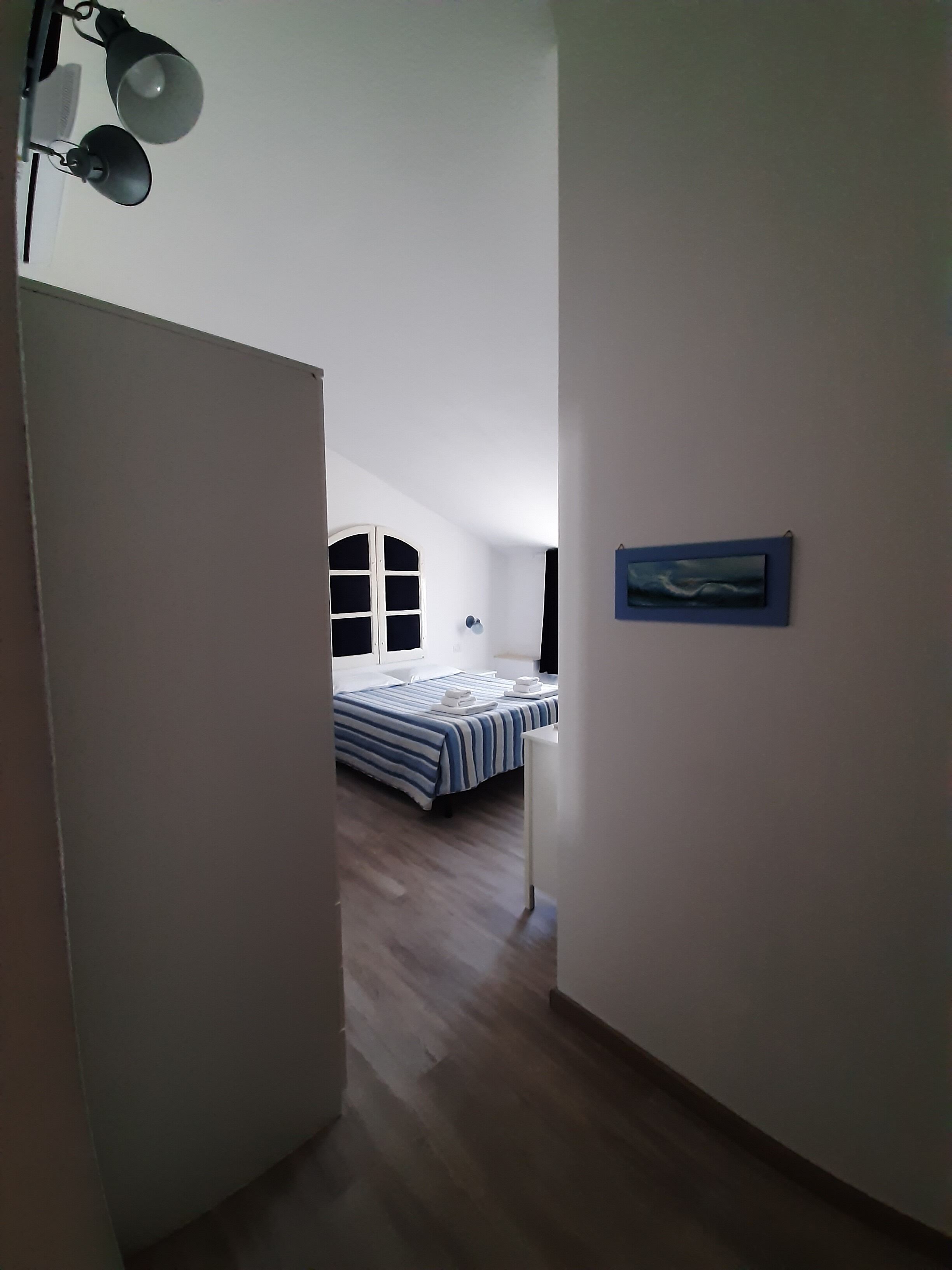 Ein schmaler Flur führt zu einem Schlafzimmer mit einem Bett, einem Fenster und einem Bild an der Wand.