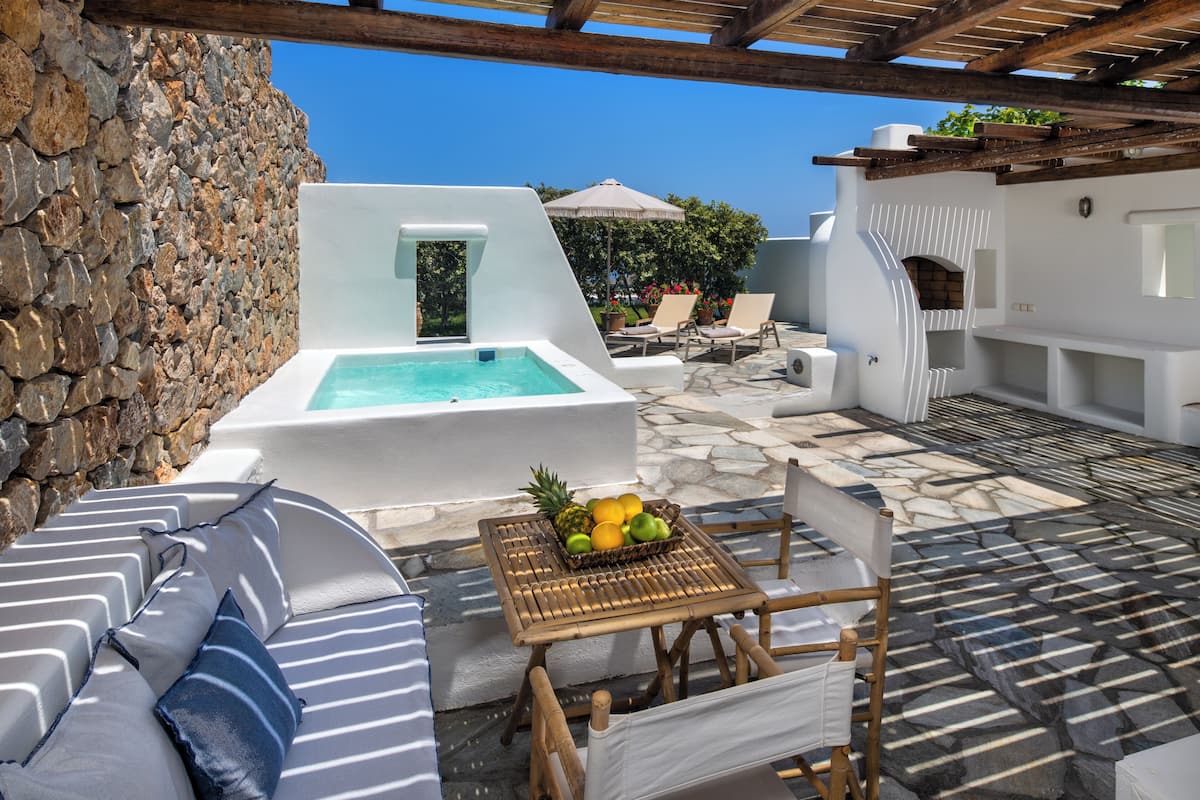 A poolside area with a stone wall, lounge chairs, a table with fruit, and a white building with a fireplace.