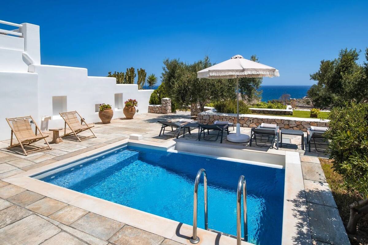 A swimming pool with lounge chairs and umbrellas, surrounded by white walls and greenery, with a view of the sea.
