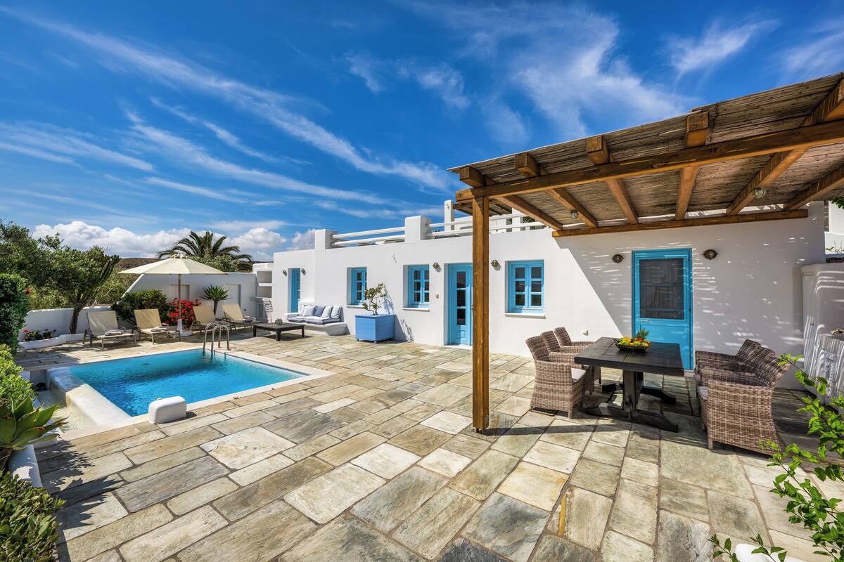 A pool area with stone flooring, white buildings with blue accents, and outdoor furniture.