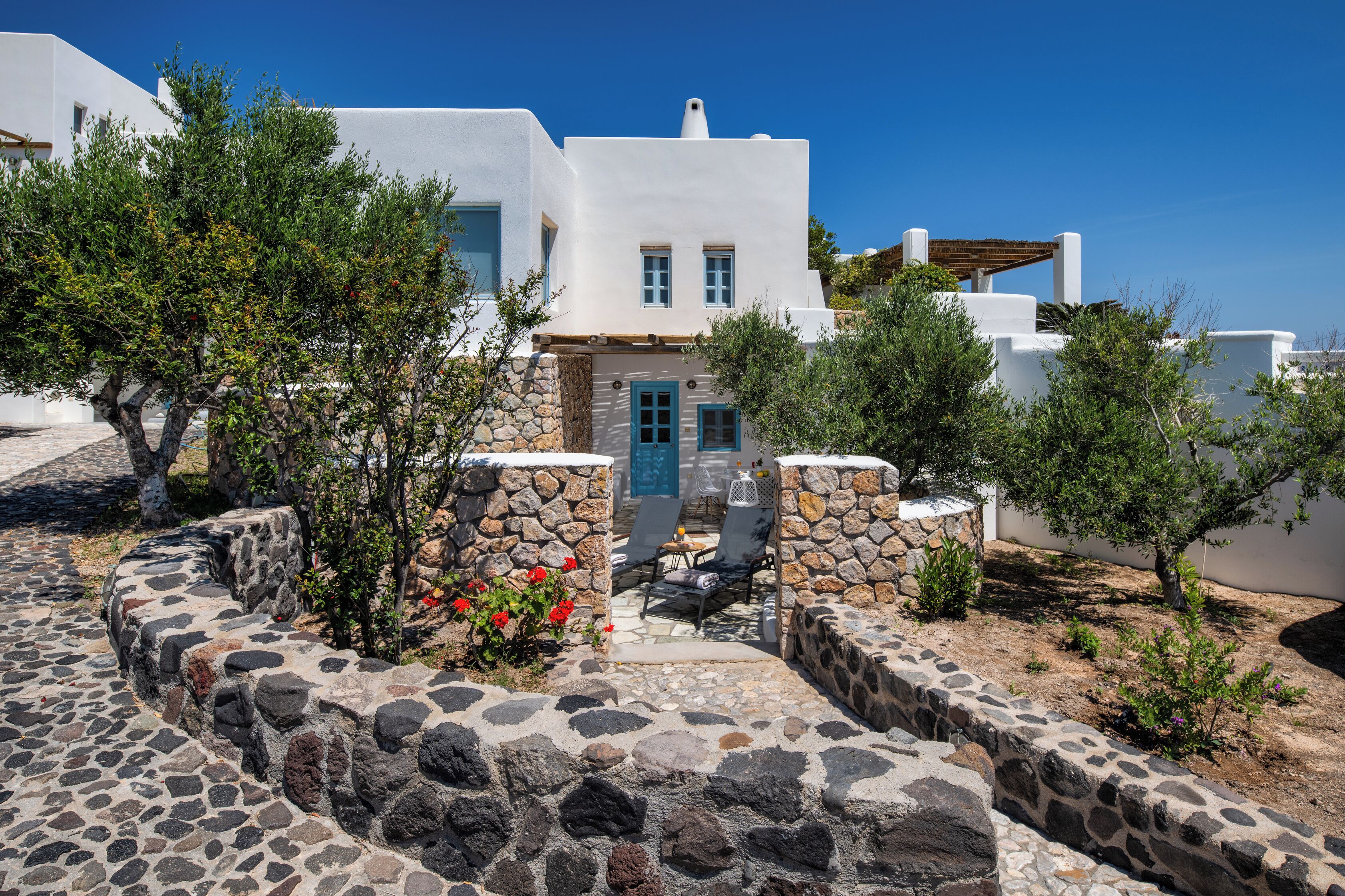 Un edificio blanco con un sendero de piedra que conduce a una puerta, rodeado de vegetación y un cielo azul despejado.