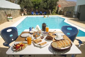 Une table de petit-déjeuner installée au bord de la piscine, avec une variété de plats et de boissons, entourée de chaises et offrant une vue sur la piscine et les clients.