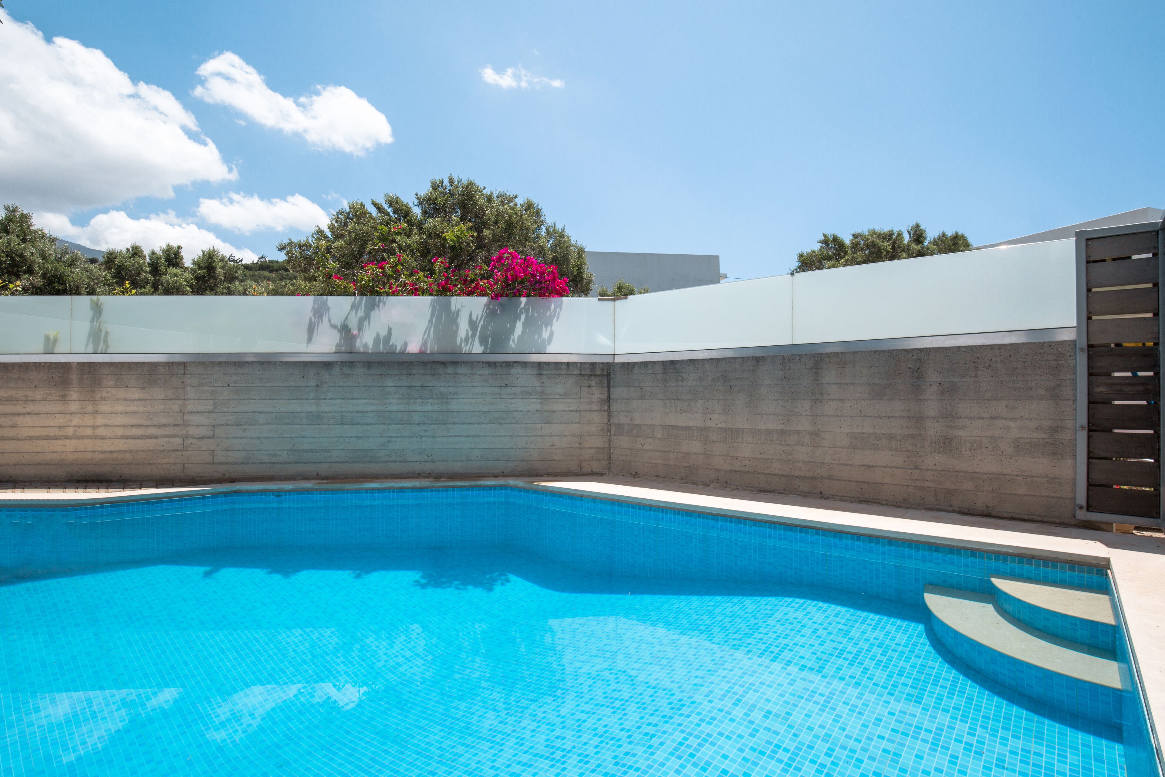 A swimming pool with clear blue water, surrounded by a concrete wall and a glass fence.