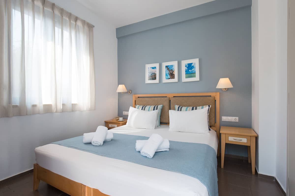 Une chambre d’hôtel avec un grand lit, deux tables de chevet, une petite table et une fenêtre avec des rideaux légers.