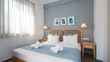 Une chambre d’hôtel avec un grand lit, deux tables de chevet, une petite table et une fenêtre avec des rideaux légers.