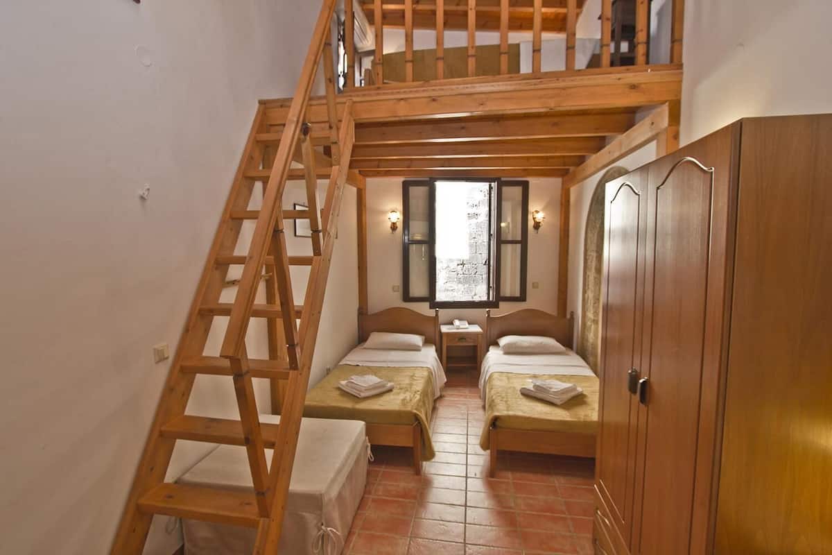 Une chambre avec deux lits, une échelle en bois, une armoire et une fenêtre donnant sur un mur de pierre.