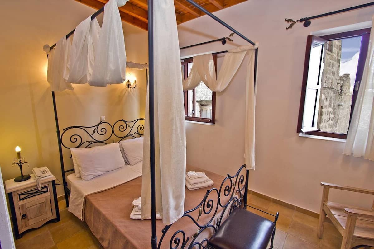 Un lit à baldaquin avec du linge de lit blanc, une table de chevet avec une lampe et une fenêtre donnant sur un bâtiment en pierre.