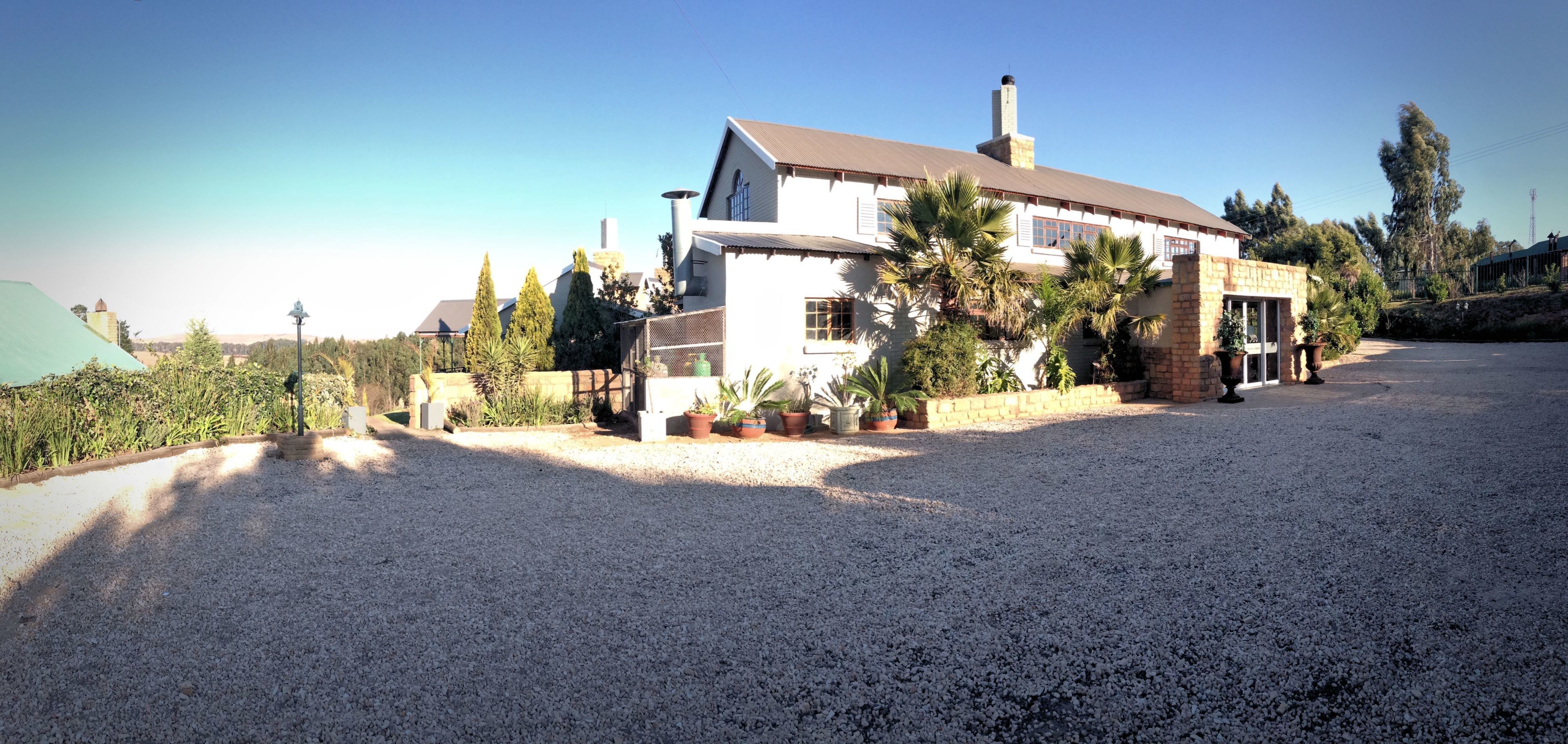 A two-story house with a gravel driveway, surrounded by potted plants and trees.