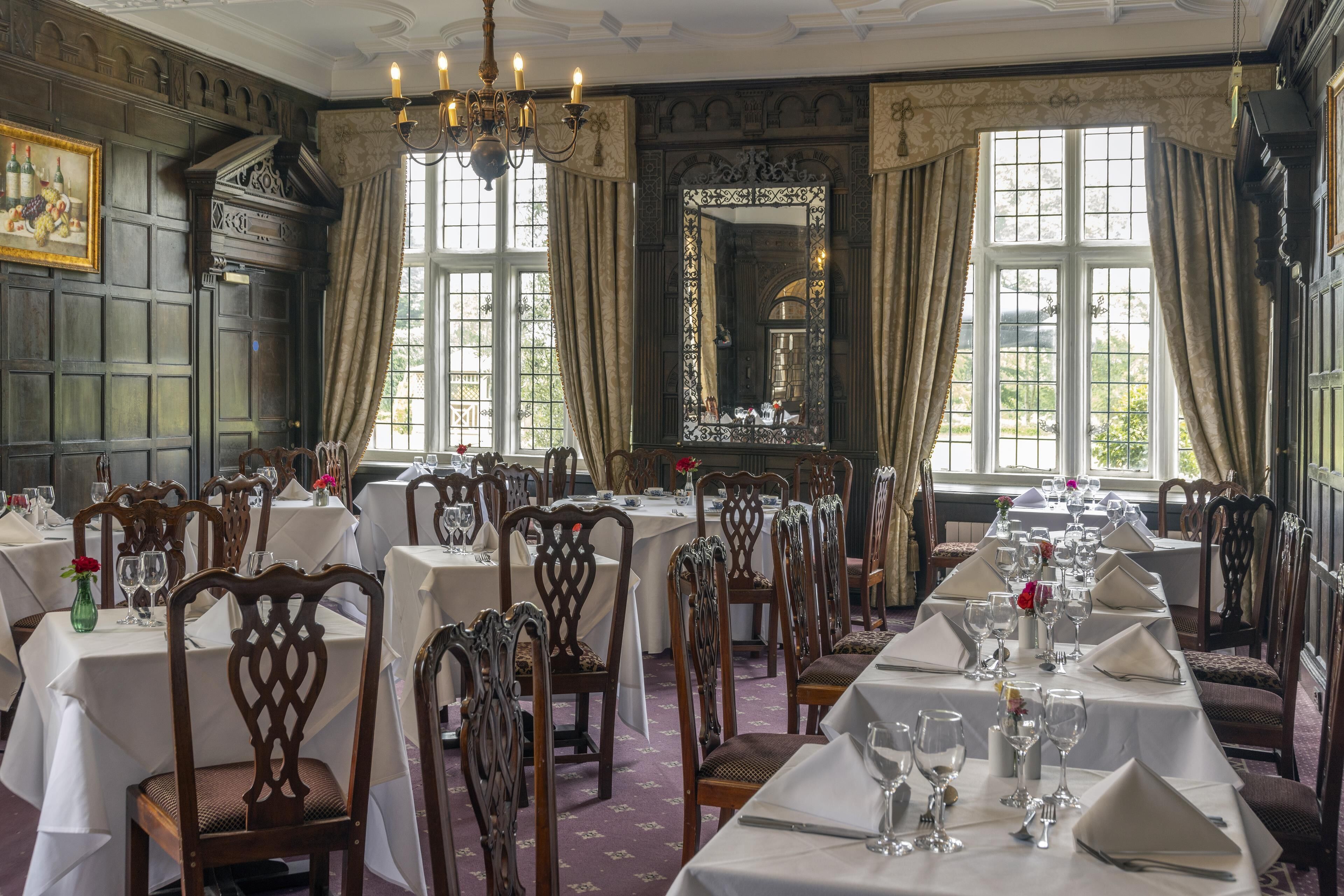 A formal dining room with multiple tables set for service, elegant wooden chairs, and large windows with curtains.