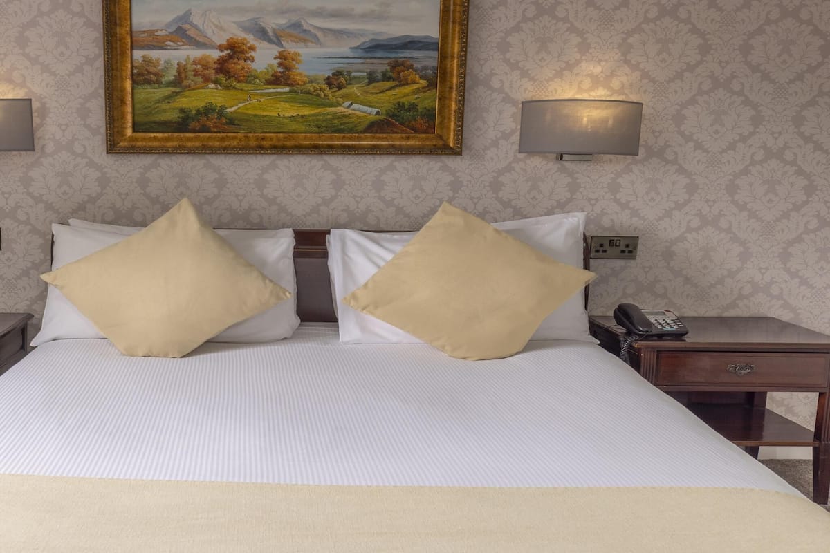 A hotel room with a large bed, two bedside tables, a painting on the wall, and two wall-mounted lamps.