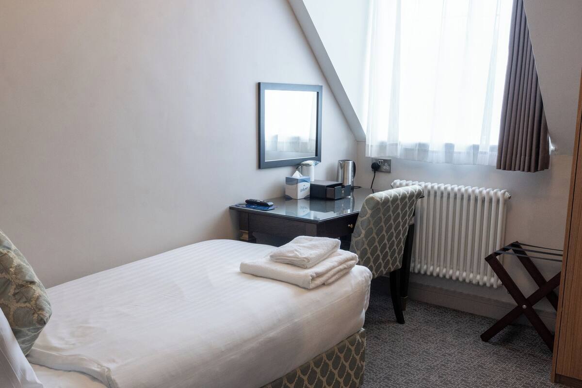 A small, modern hotel room with a bed, a desk, a chair, a radiator, and a window with curtains.