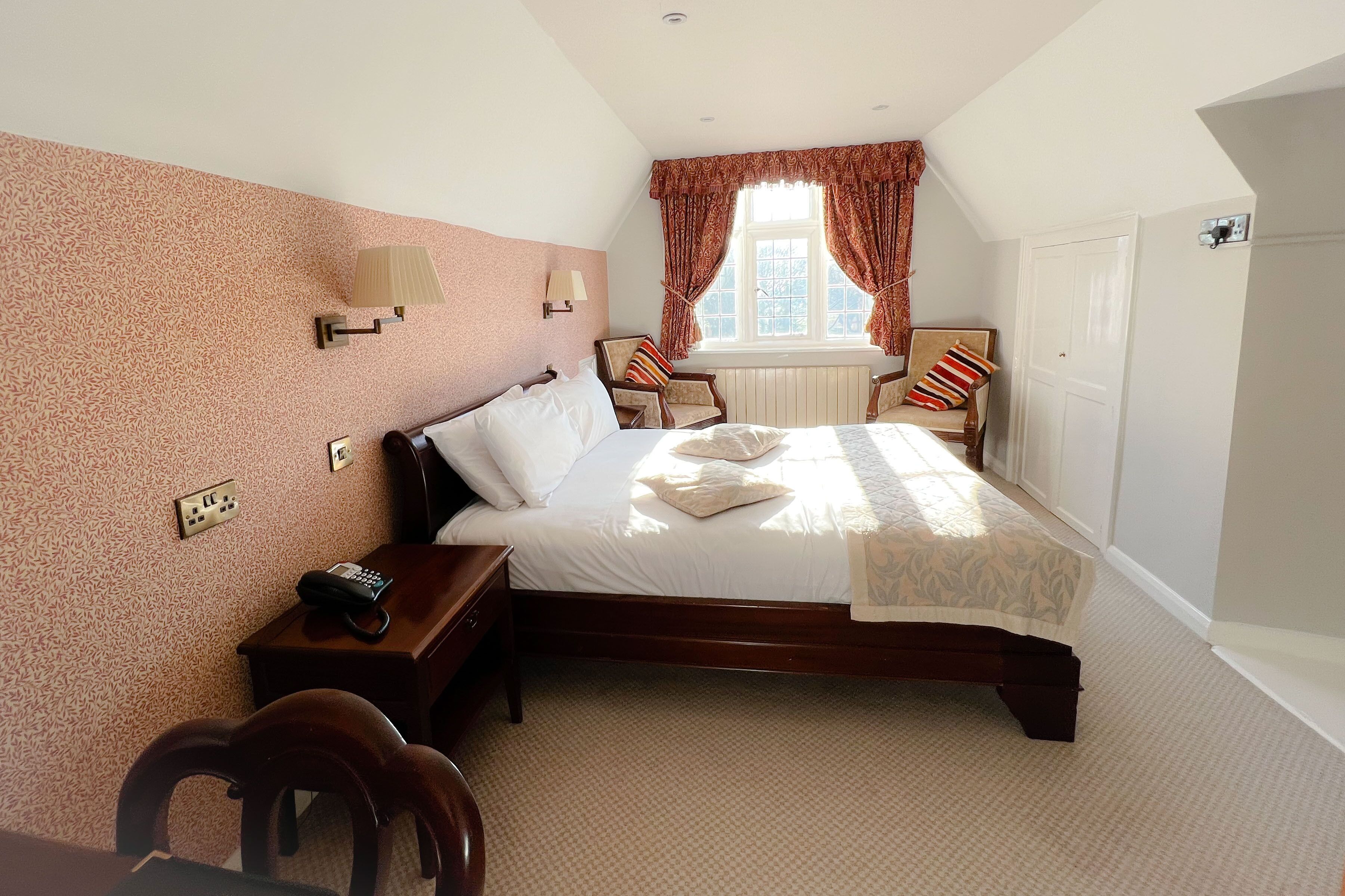 A bedroom with a large bed, a nightstand, a chair, a window with curtains, and a wall-mounted lamp.