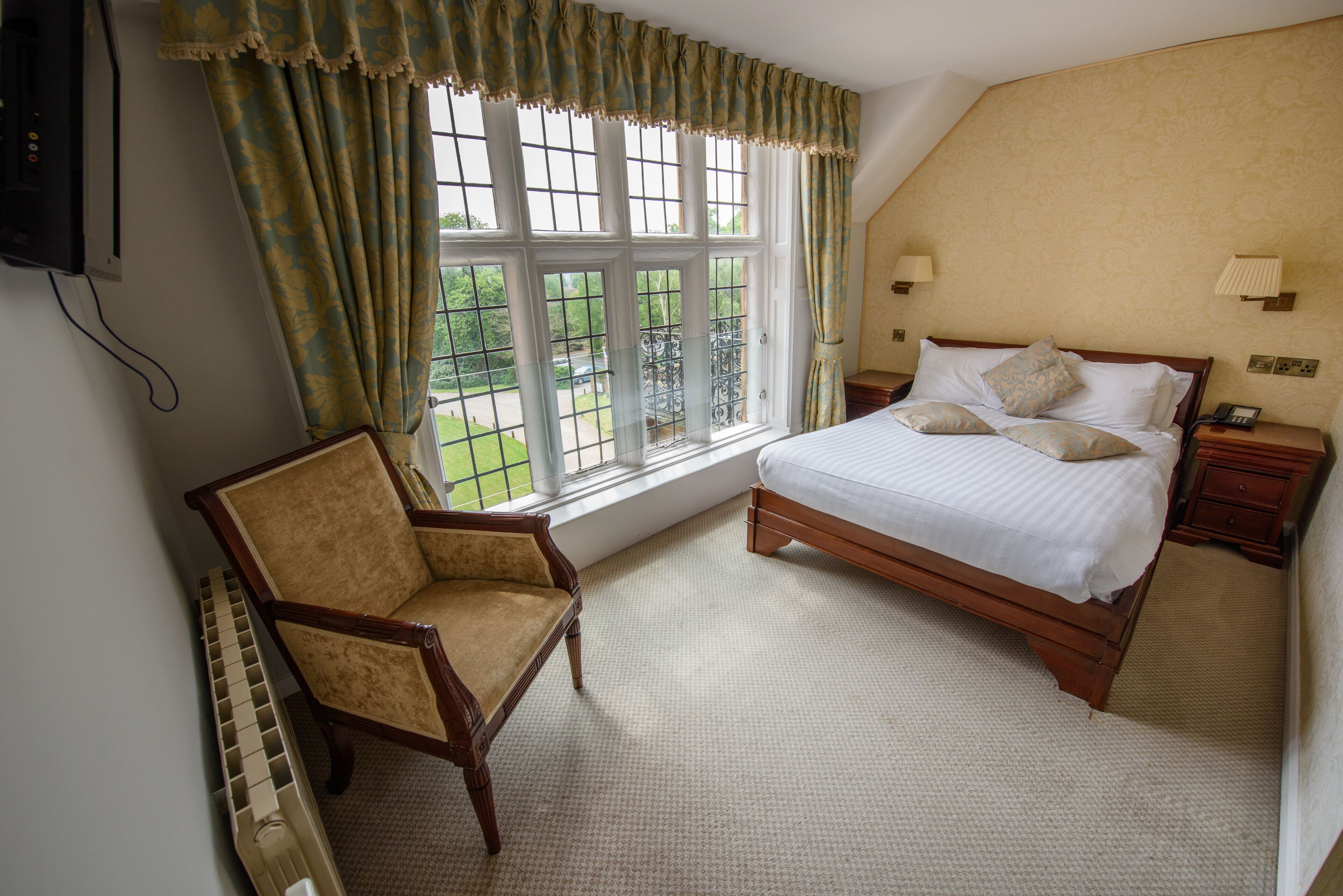 A bedroom with a large bed, a chair, a nightstand, a lamp, and a window with curtains.