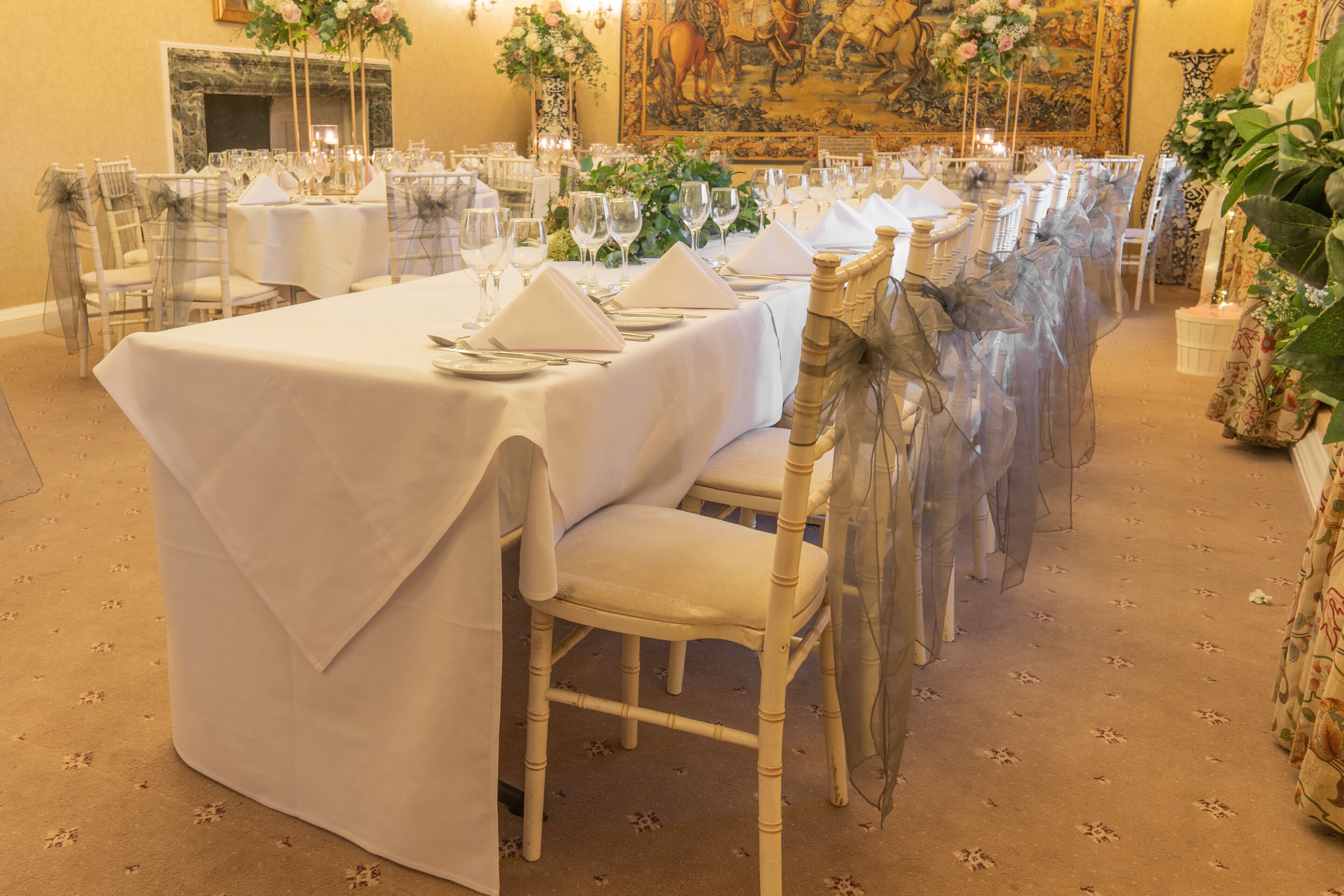 Un salón de banquetes con largas mesas dispuestas para un evento formal, con manteles blancos, sillas transparentes y centros de mesa florales.
