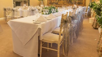 A banquet hall with long tables set for a formal event, featuring white tablecloths, clear chairs, and floral centerpieces.