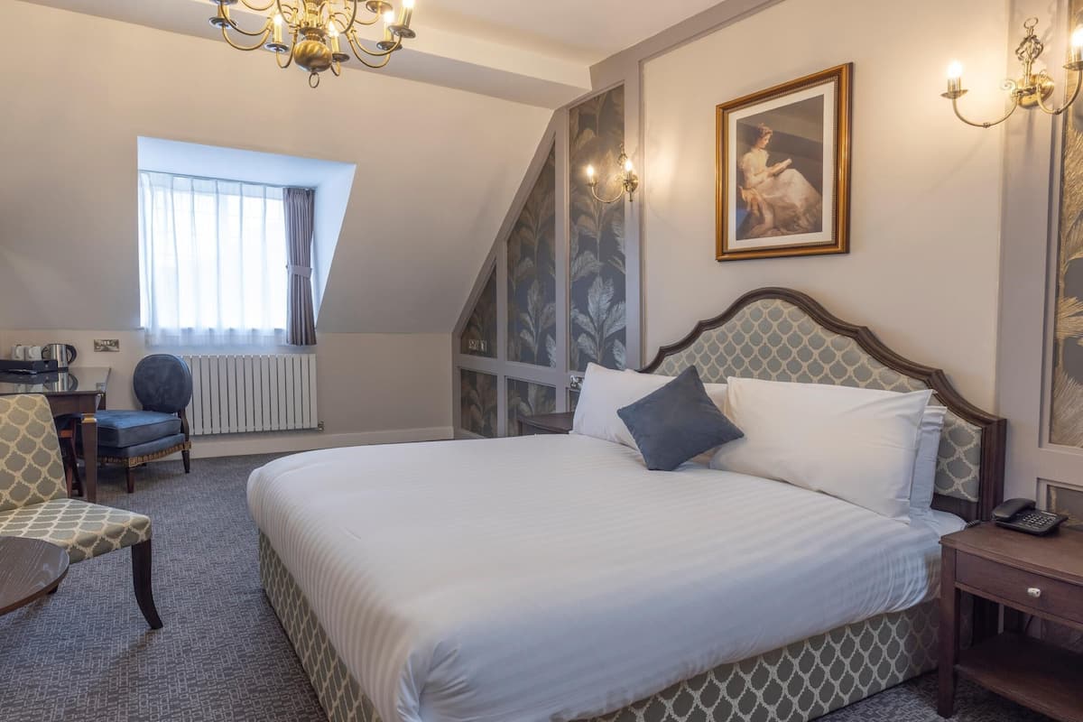 A hotel room with a bed, a desk, a chair, a small table, a radiator, a chandelier, and a painting on the wall.