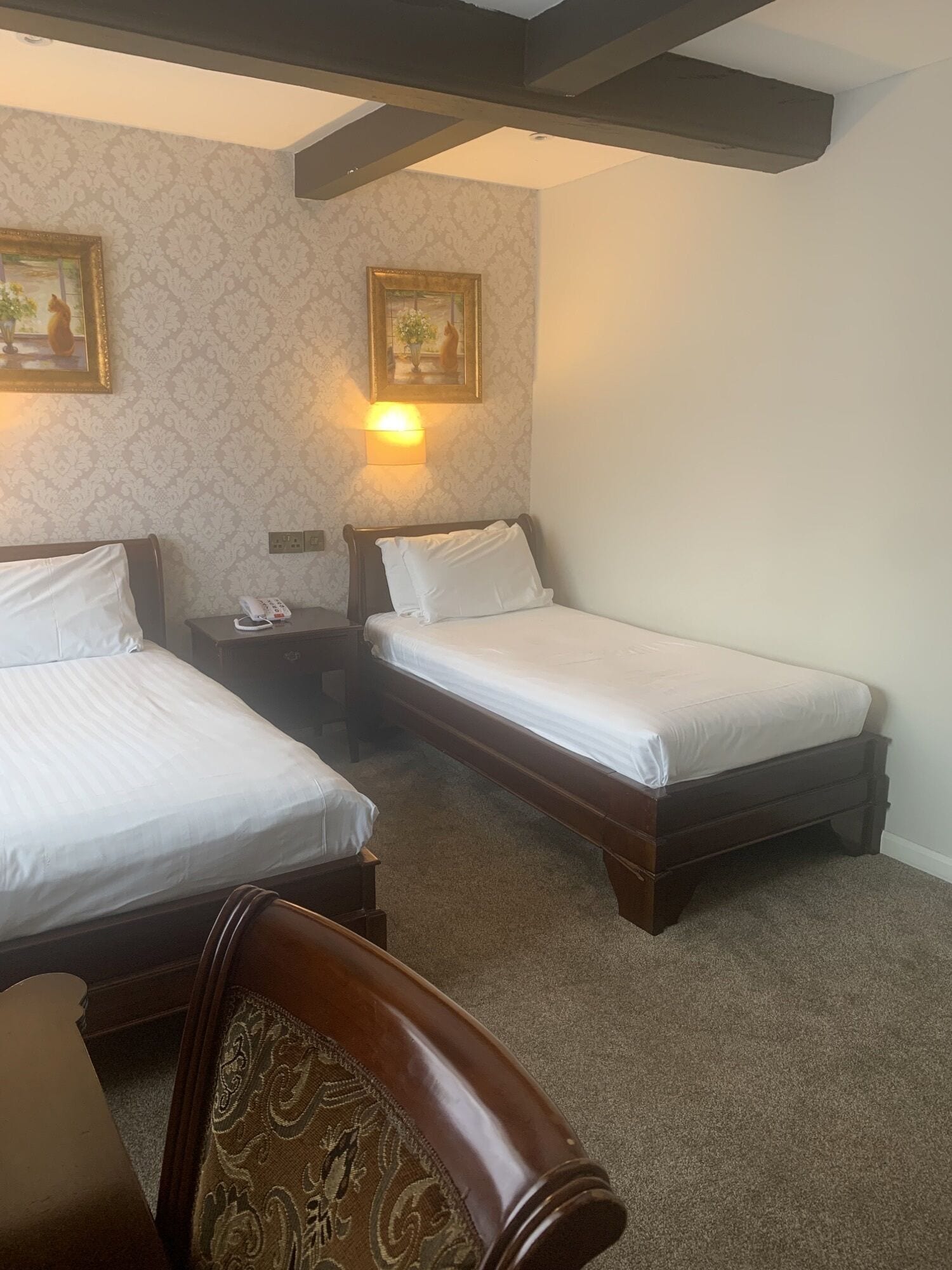 A hotel room with two single beds, a wooden chair, a nightstand with a phone, and a wall-mounted lamp.