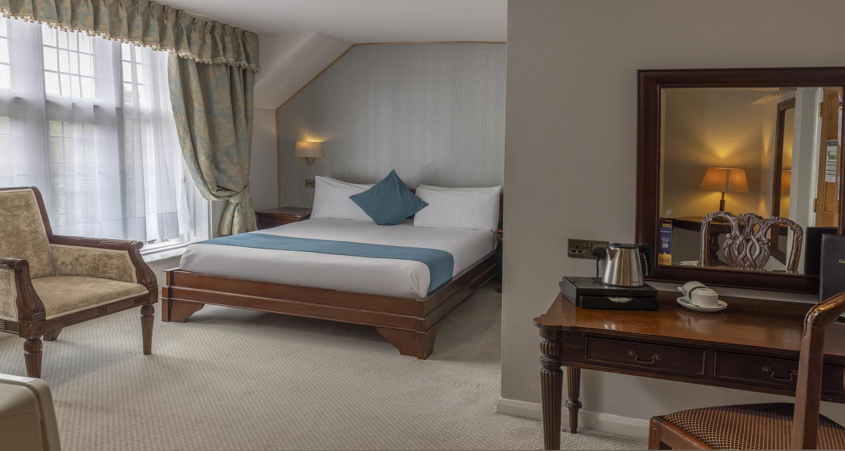 A hotel room with a bed, a chair, a desk with a mirror, and a lamp.