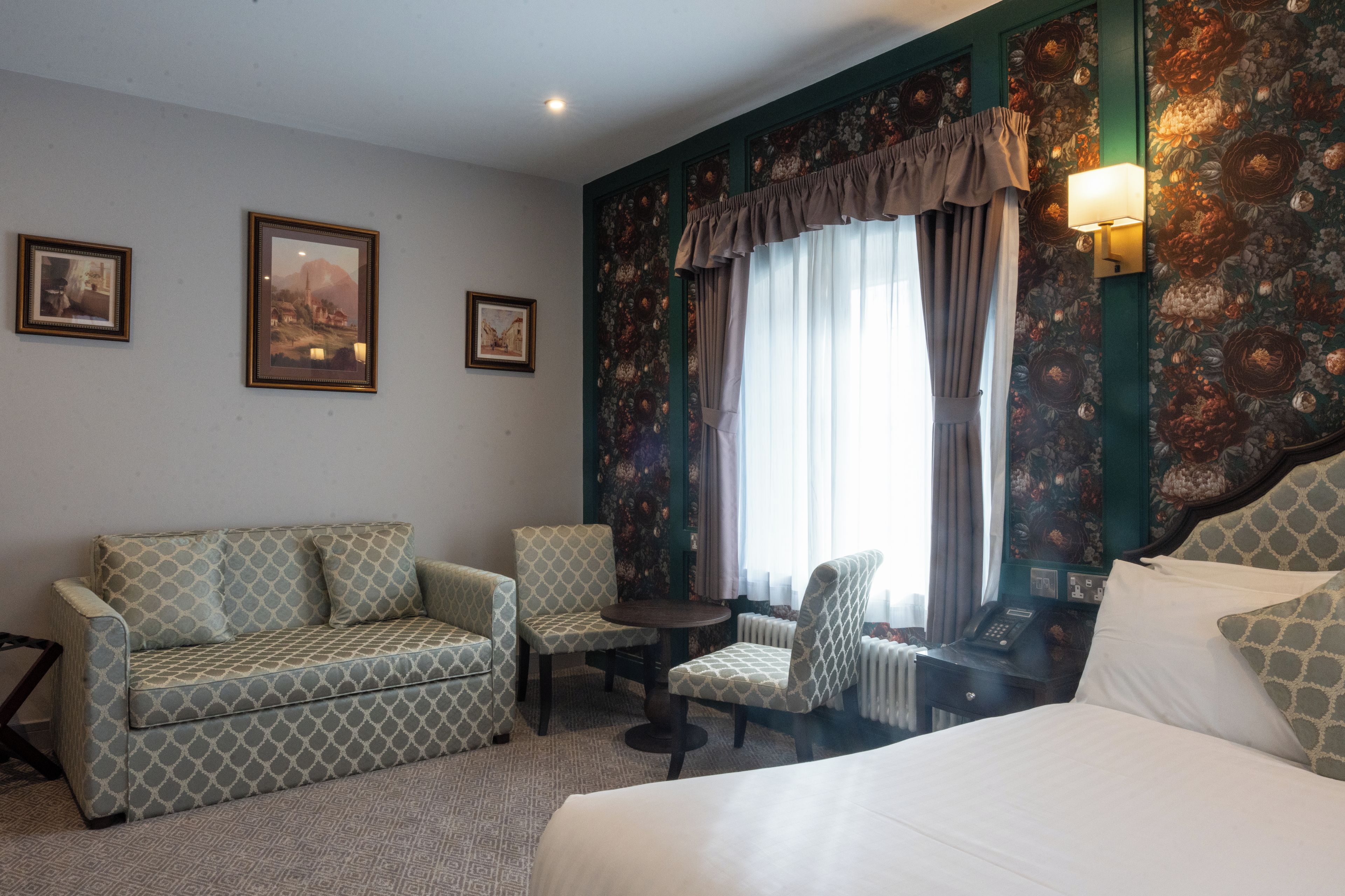 Quarto de hotel com uma cama, duas poltronas, uma mesinha e duas imagens emolduradas na parede.