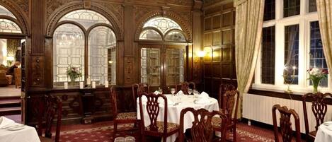 A formal dining room with ornate wood paneling, large arched windows, and chandeliers.