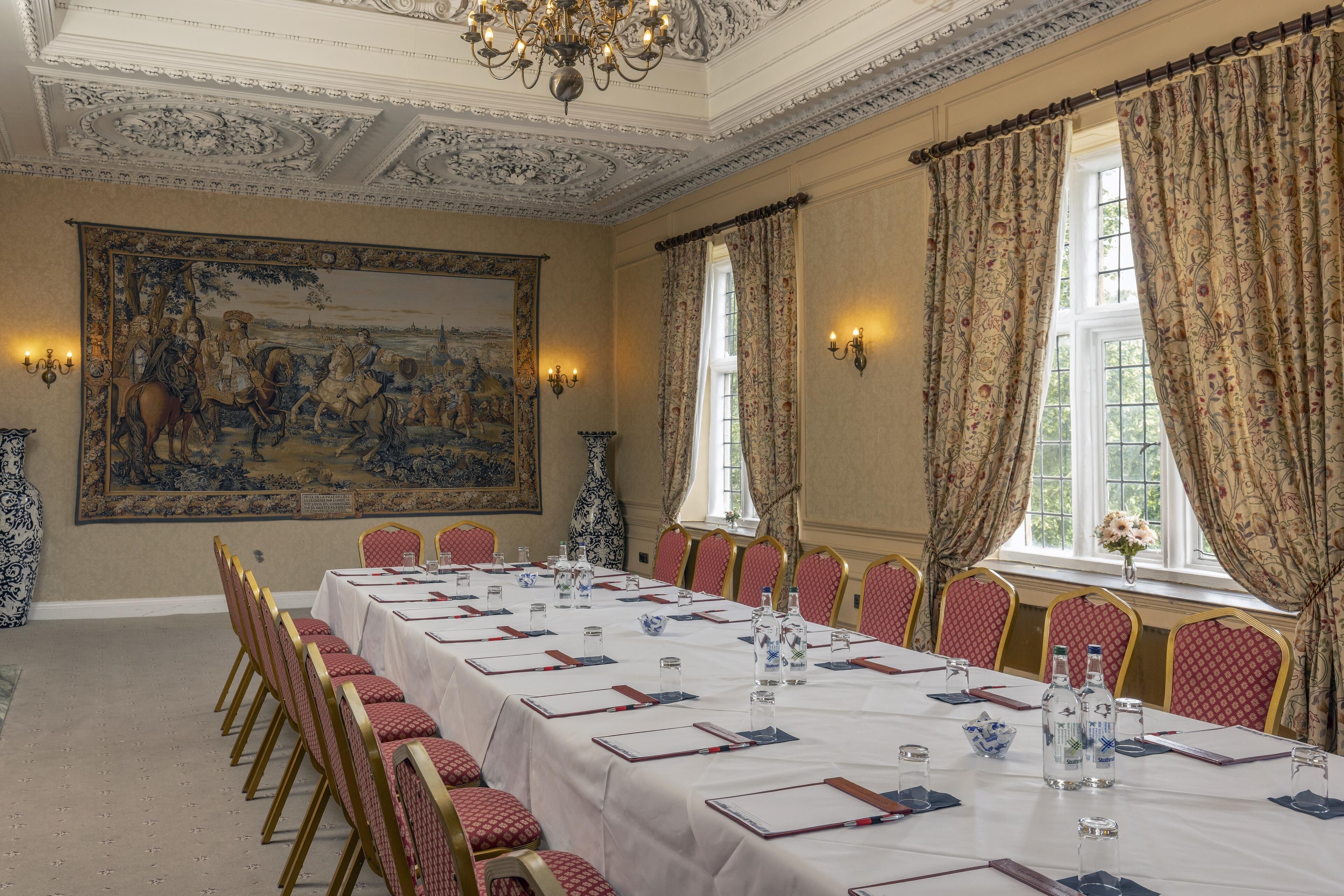 A formal dining room with a long table set for a meal, red upholstered chairs, and a large historical painting on the wall.