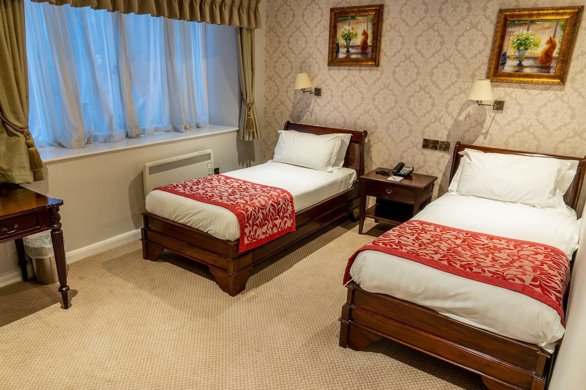A hotel room with two wooden beds, each with a red and white bedspread, a window with curtains, a radiator, a nightstand with a lamp, and a framed painting on the wall.