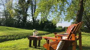 Una silla Adirondack de madera y una mesita con una copa de vino y una maceta en un entorno exterior con césped.