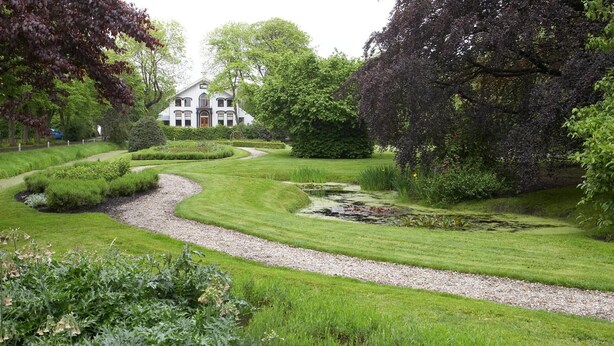 Un jardín bien cuidado con un sendero serpenteante, un estanque y una casa grande al fondo.