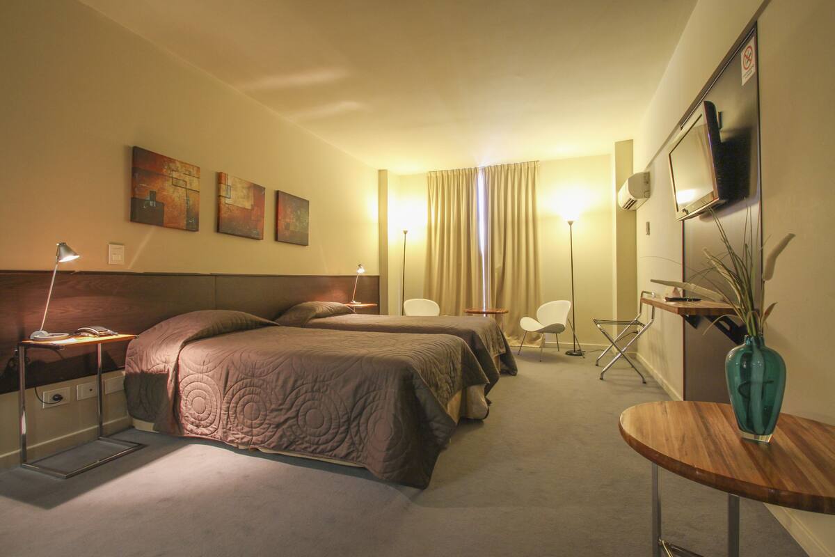 A hotel room with two beds, a desk, a chair, a TV, and artwork on the wall.