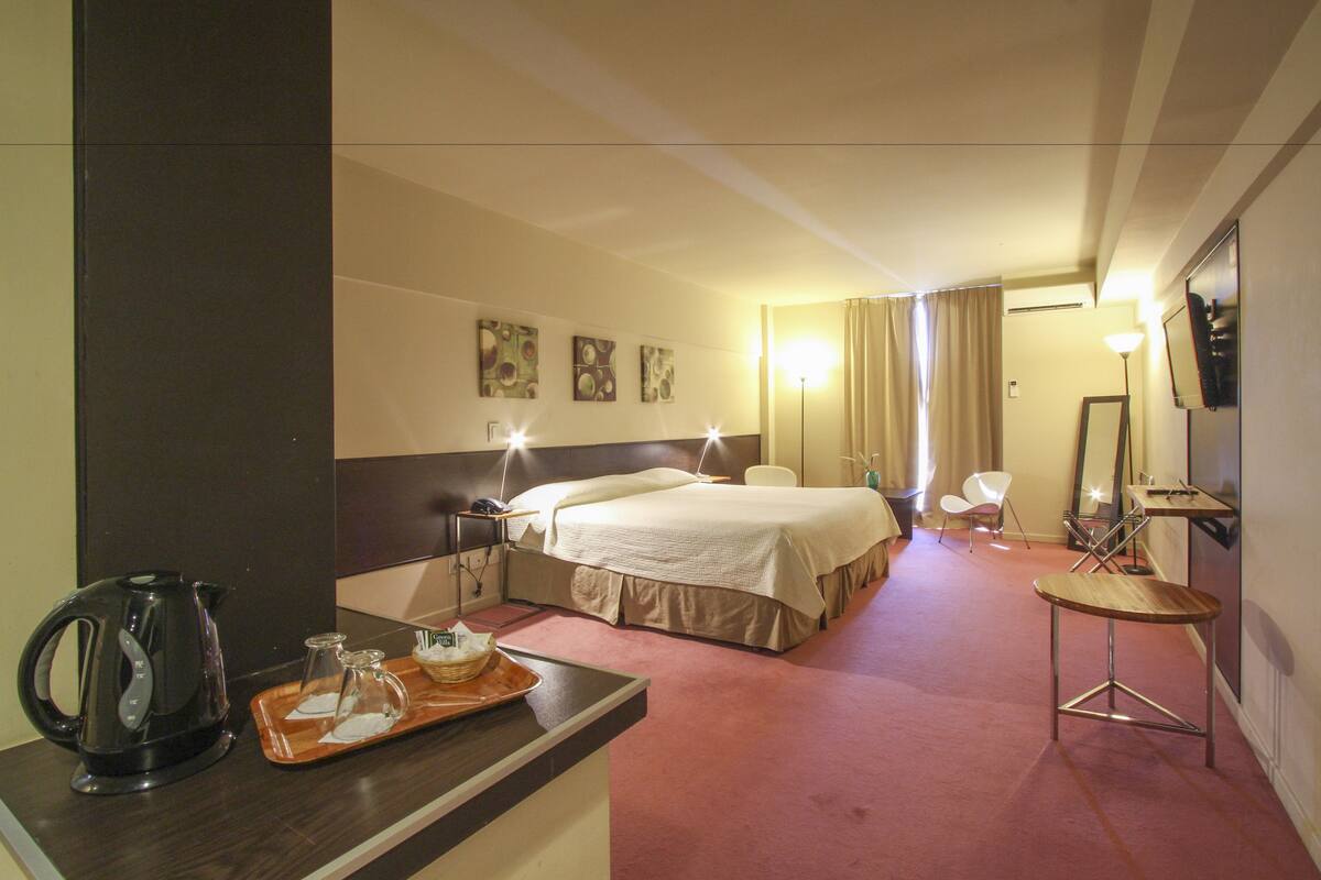 A hotel room with a bed, bedside table, lamp, chair, and a small table with a tray of breakfast items.
