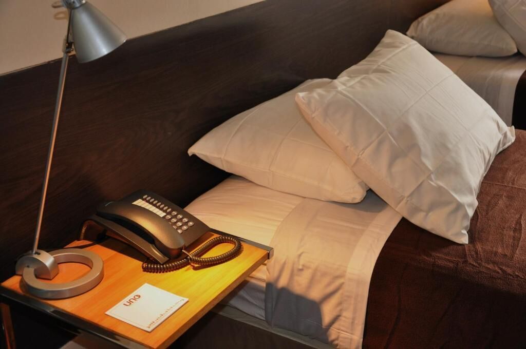 Habitación de hotel con cama, teléfono de noche, lámpara y una libreta con la palabra 'UNO' escrita.