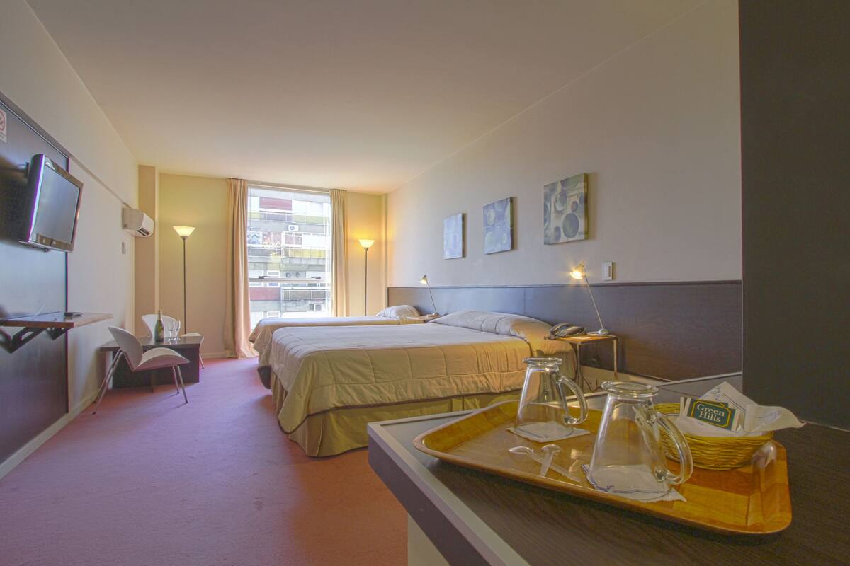 A hotel room with two beds, a TV, a desk with a tray of breakfast items, and a window with a view of buildings.
