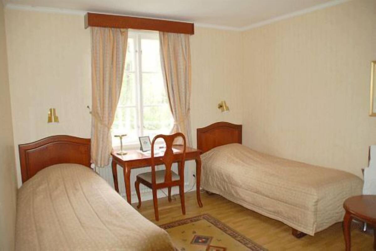Una habitación con dos camas individuales, un escritorio de madera con una silla, una ventana con cortinas y una mesita pequeña.