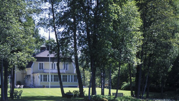 A lakeside view of a large, yellow building with a balcony, surrounded by trees and a grassy area with outdoor furniture. - Solvikens Pensionat (Ingelstad)