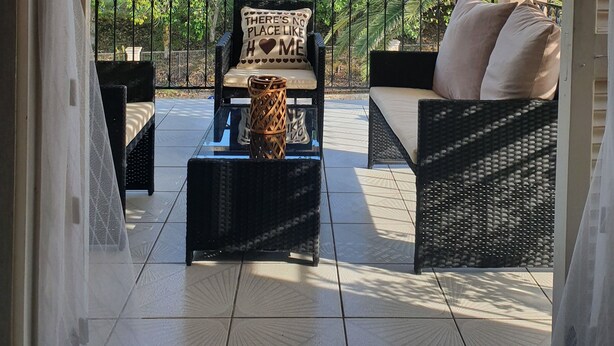 A covered outdoor seating area with wicker furniture, a glass coffee table, and a view of lush greenery and a building in the background. - Asprokavos Beach Apartment (Corfu)