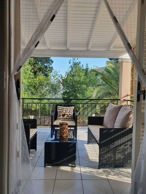 A covered outdoor seating area with wicker furniture, a glass coffee table, and a view of lush greenery and a building in the background. - Asprokavos Beach Apartment (Corfu)