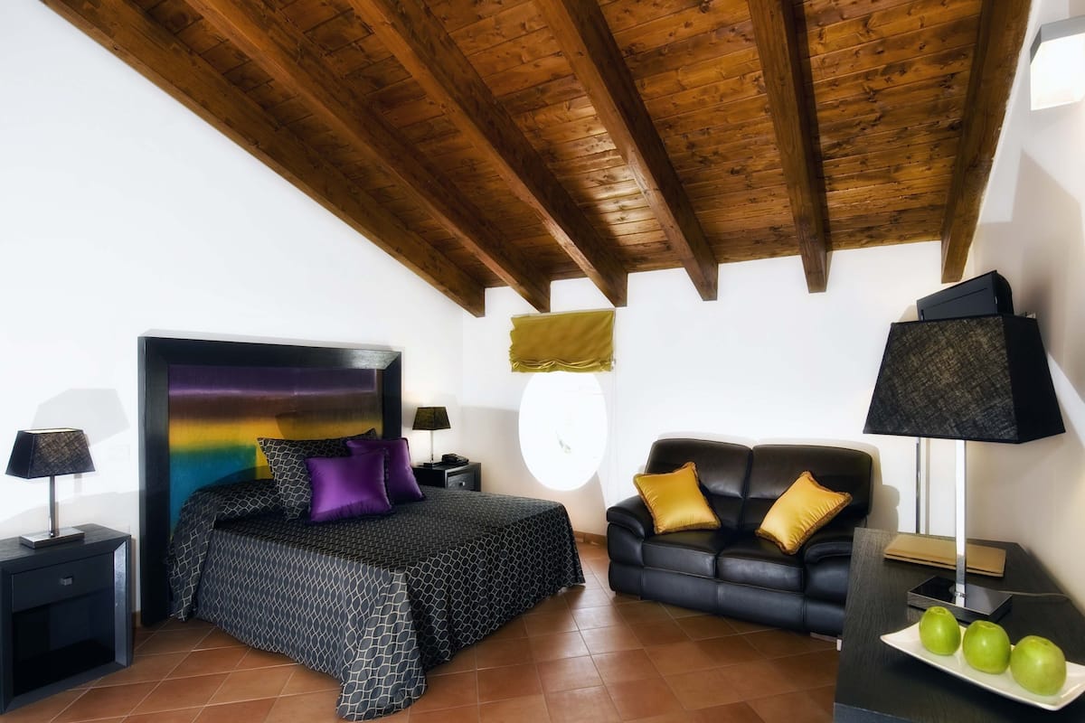 A bedroom with a bed, a black leather sofa, a nightstand, a lamp, and a window with a view.