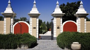 Una entrada imponente con dos pilares blancos y puertas rojas, que da a un camino bordeado por setos cuidados.