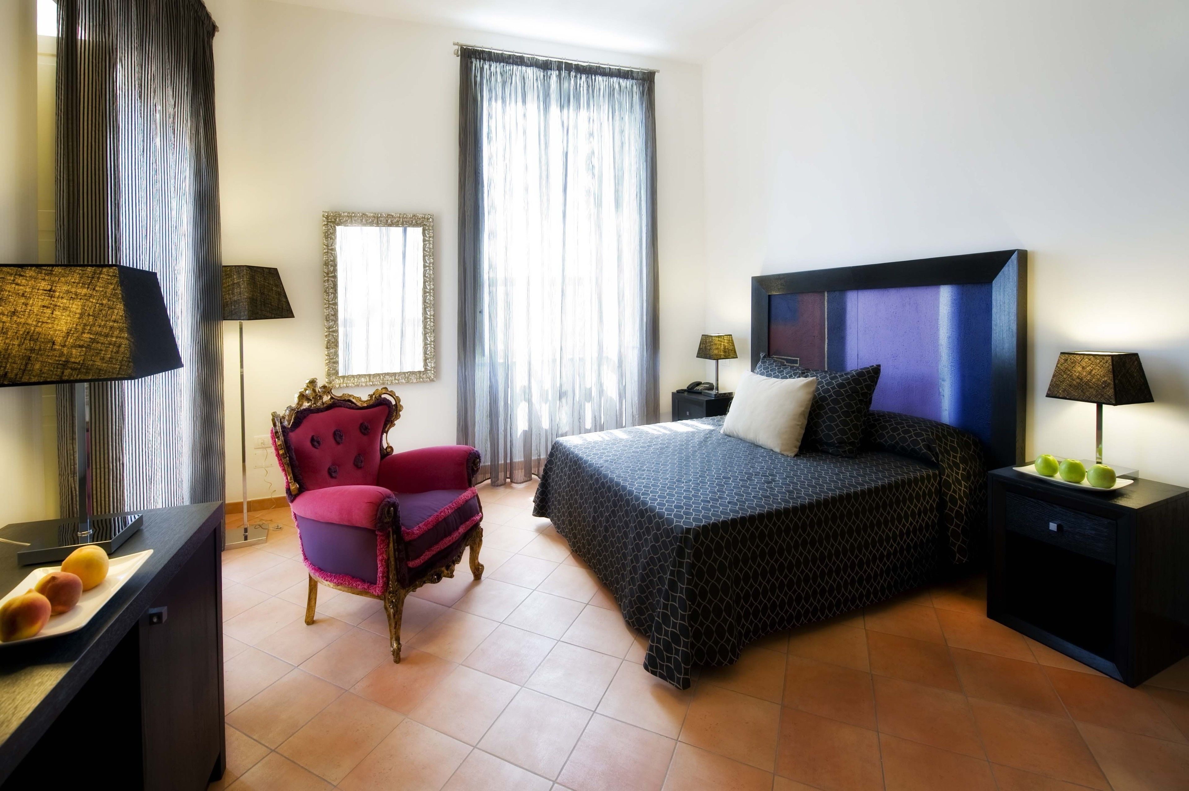 Habitación de hotel con cama, una silla, un escritorio con lámpara y una mesita de noche con lámpara.